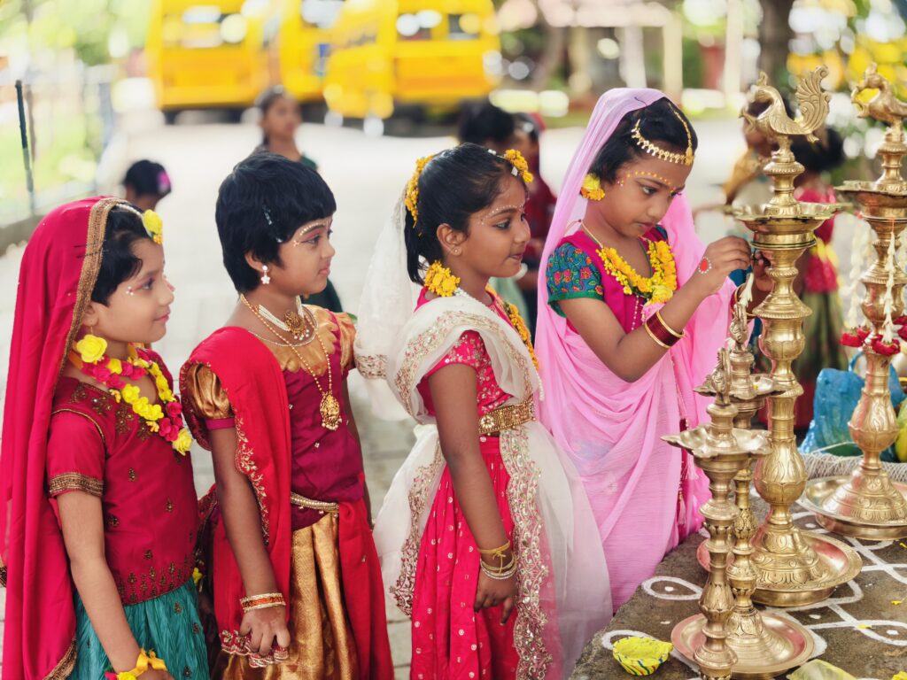 Children in festive costumes watch as one child reaches towards a tall brass lamp.