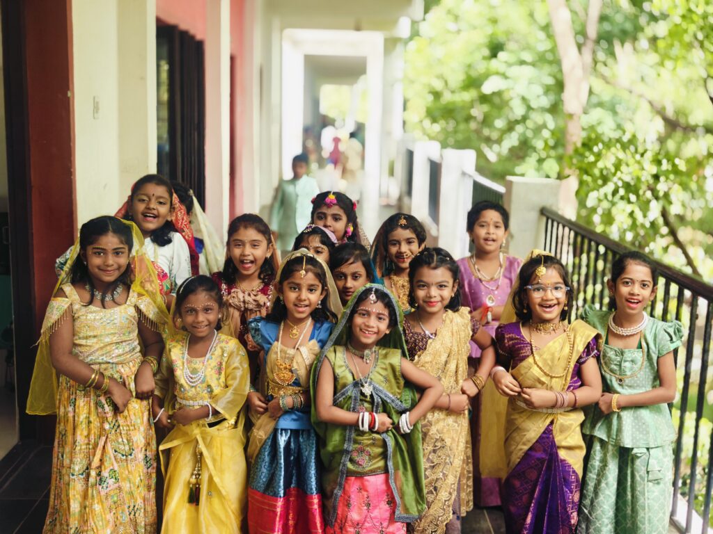 A group of smiling children in traditional outfits pose together in a school corridor.