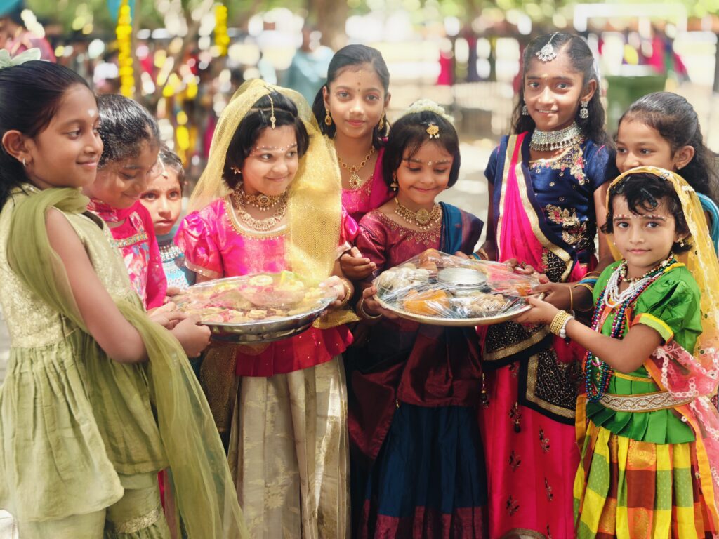 A group of children in traditional dress hold two covered plates of food and smile at the camera.