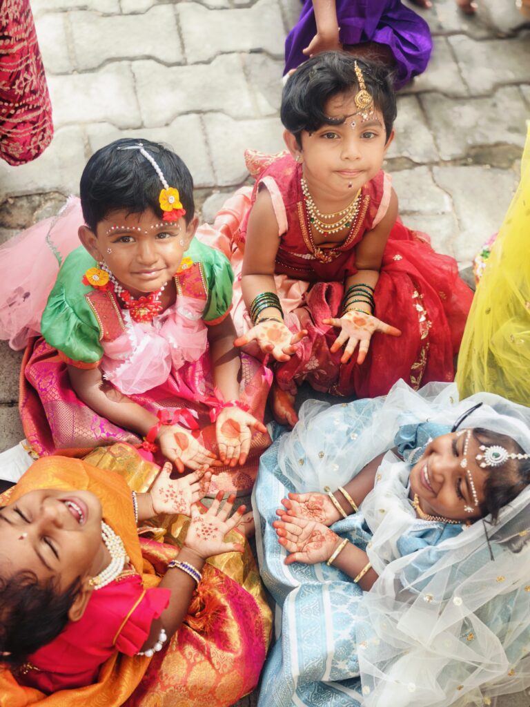 Four young children in festive costumes sit on the ground showing the henna on their palms.