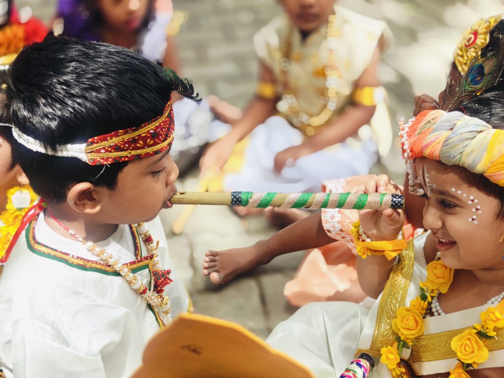 A child dressed as Krishna blows a decorated flute held by another child in a colourful turban and flower garland.