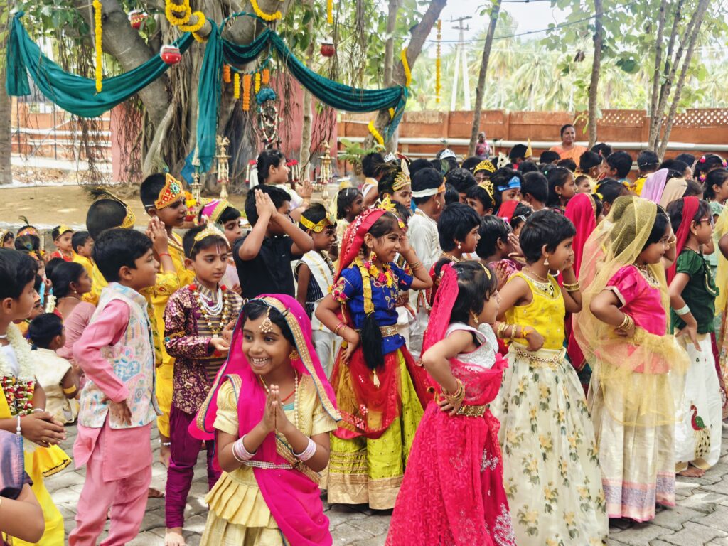 A large group of children in costume dance in a courtyard beside a decorated banyan tree.