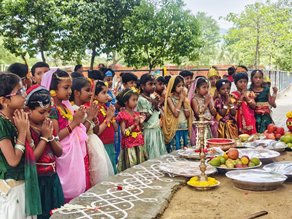 Children in traditional clothes stand with folded hands before a brass lamp, fruit and kolam on a ledge.