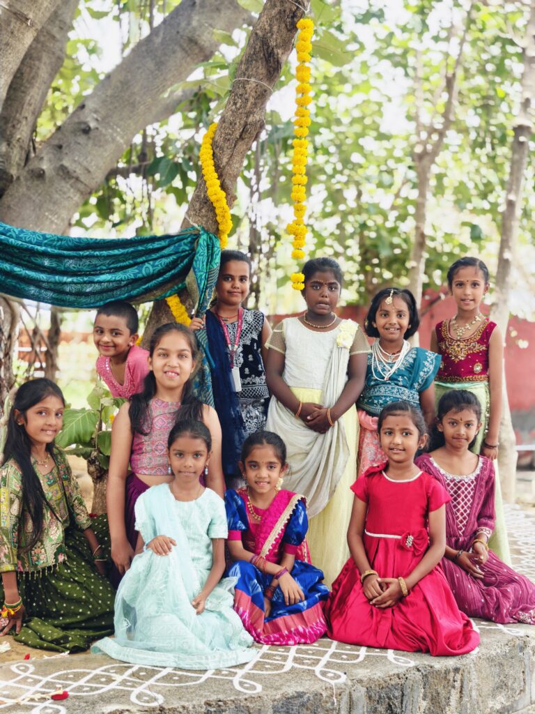 Children in festive outfits sit and stand together under a tree hung with marigolds and teal cloth.