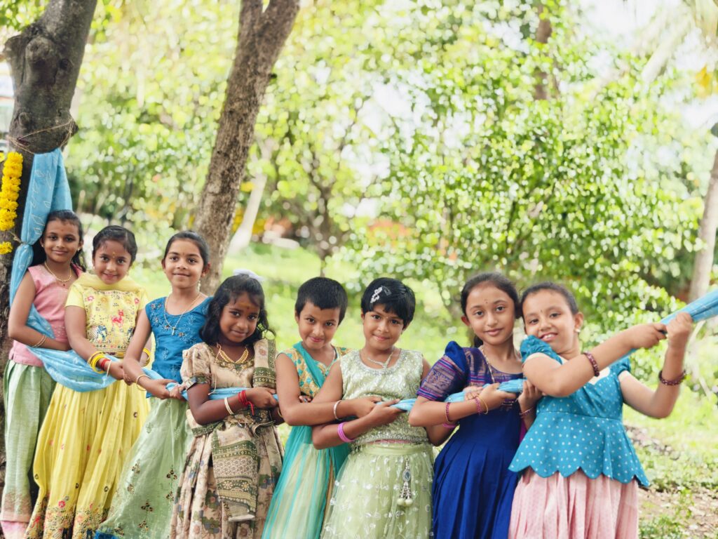 Children in festive dresses stand in a line holding a light blue cloth tied to a tree.