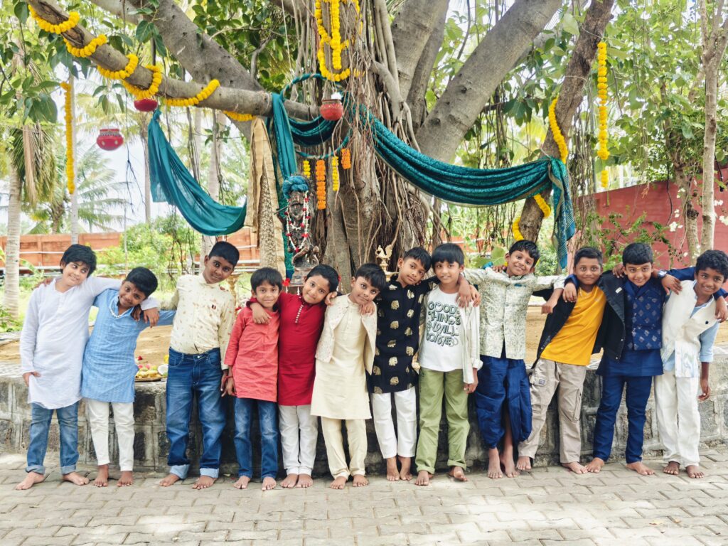 A row of boys in kurtas and casual clothes stand arm in arm before a tree decorated with marigold garlands.