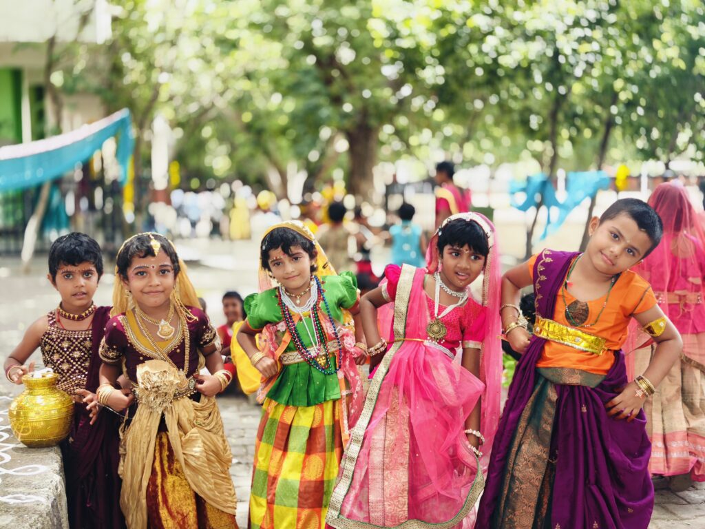 Five children in colourful traditional outfits pose outdoors, one resting a hand on a brass pot.