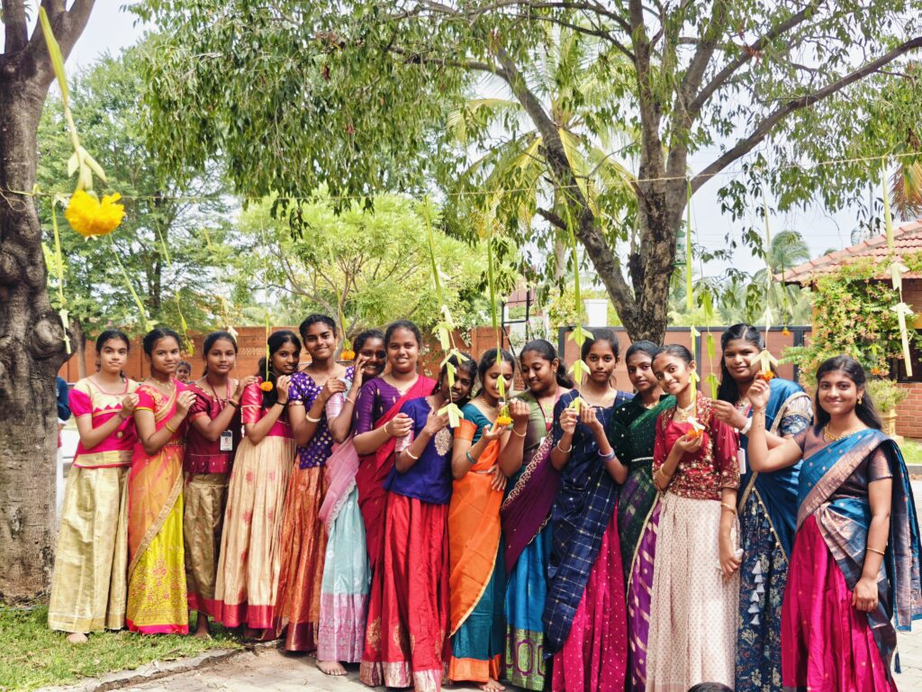 A group of girls in traditional dress hold small flowers under trees hung with decorations