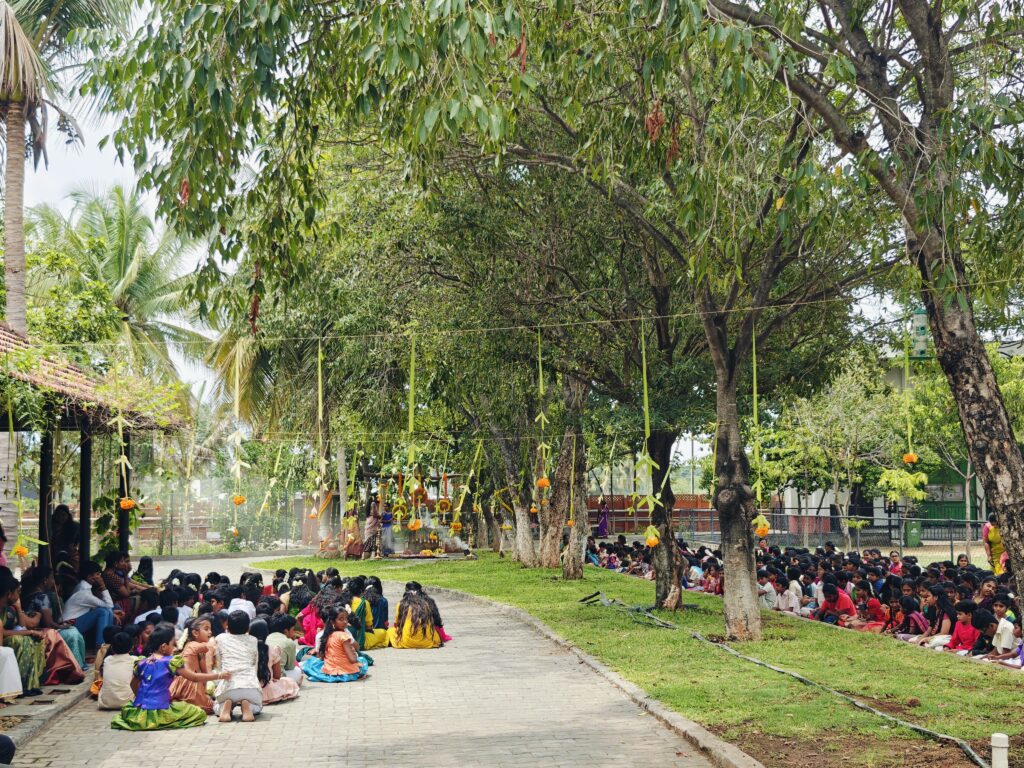 Students sit along a tree-lined path decorated with hanging flowers, facing the shrine