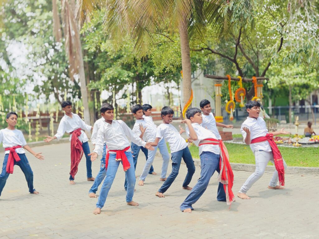 Boys in white shirts with red sashes at the waist dance on a paved courtyard under the trees