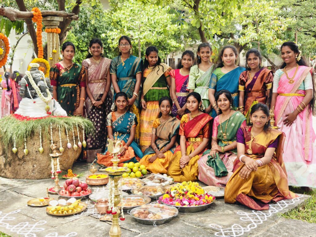 Girls in half-sarees pose in two rows beside the decorated Ganesha idol and plates of offerings