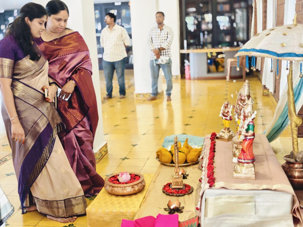 Two women in silk sarees look at a decorated display of deity figures, a brass lamp and flowers in a hall.