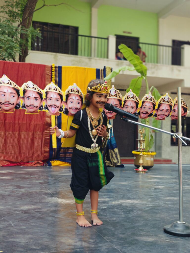 A child in a crown and painted moustache speaks into a microphone holding a row of paper face cut-outs.