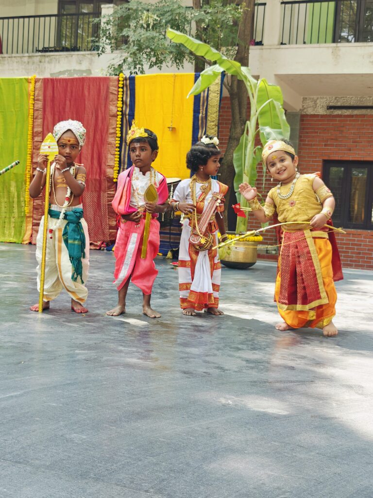 Four young children in traditional costumes stand on stage in front of a colourful saree backdrop.