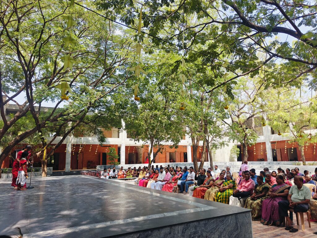 A child performs on a stage beneath trees hung with palm-leaf decorations as parents watch from chairs.