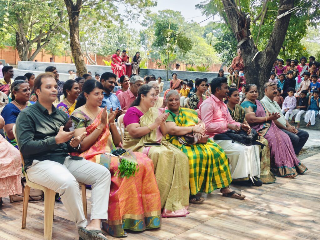 Seated parents in sarees and festive wear clap during the programme under the trees.