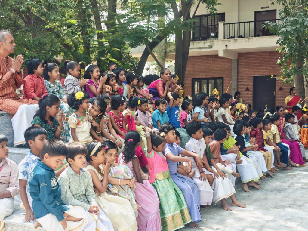 Children in festive clothes sit on courtyard steps with teachers, watching and clapping.