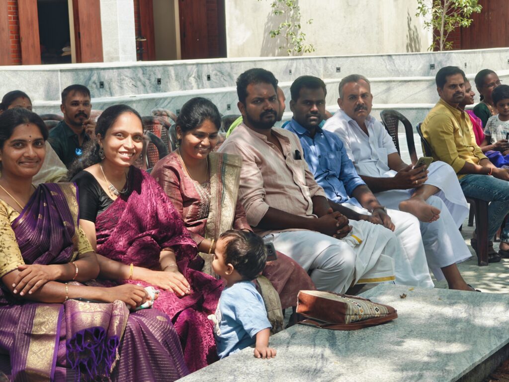 Parents sit in a row of chairs in the courtyard, smiling, as a toddler stands beside them.