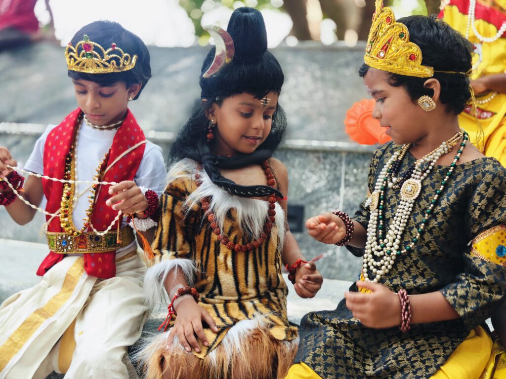 Three young children in costumes with crowns and jewellery sit together adjusting bead necklaces.