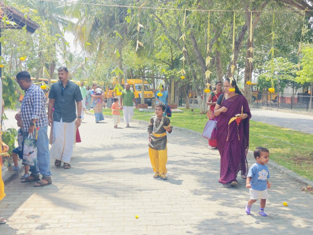 Families walk along a paved path under hanging palm-leaf and marigold decorations; a child in costume walks ahead.