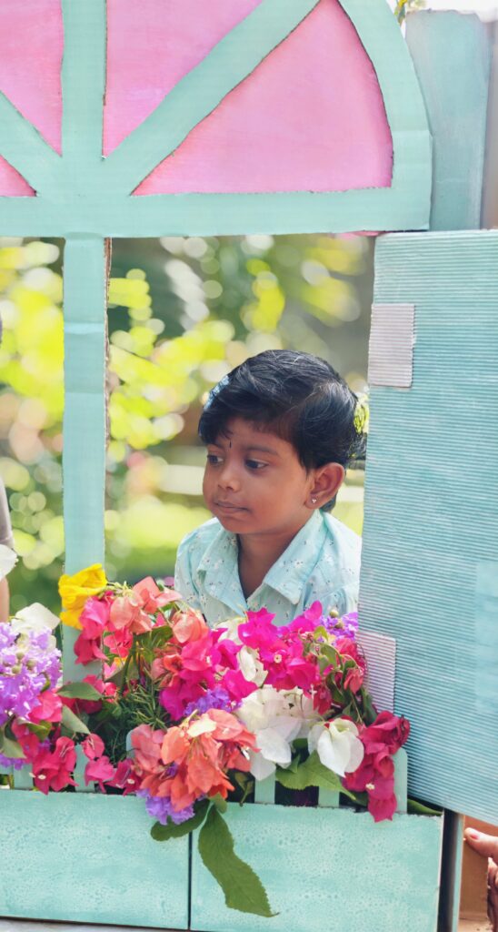 A child in a light blue top looks sideways from a cardboard window with pink and white flowers.