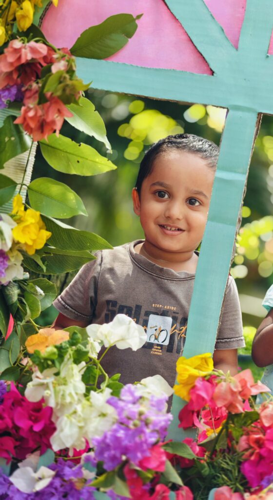 A smiling child in a grey T-shirt peeks through the window prop beside bright flowers.