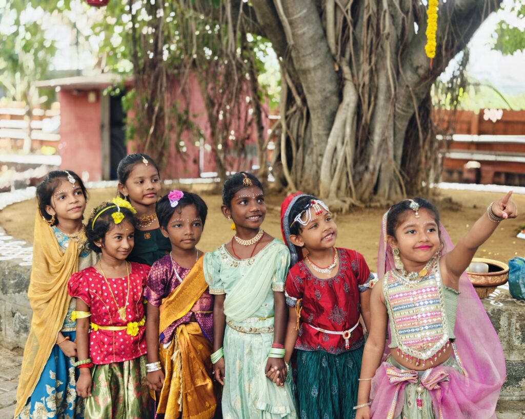 Six children in traditional dress stand by a large tree, one pointing into the distance.
