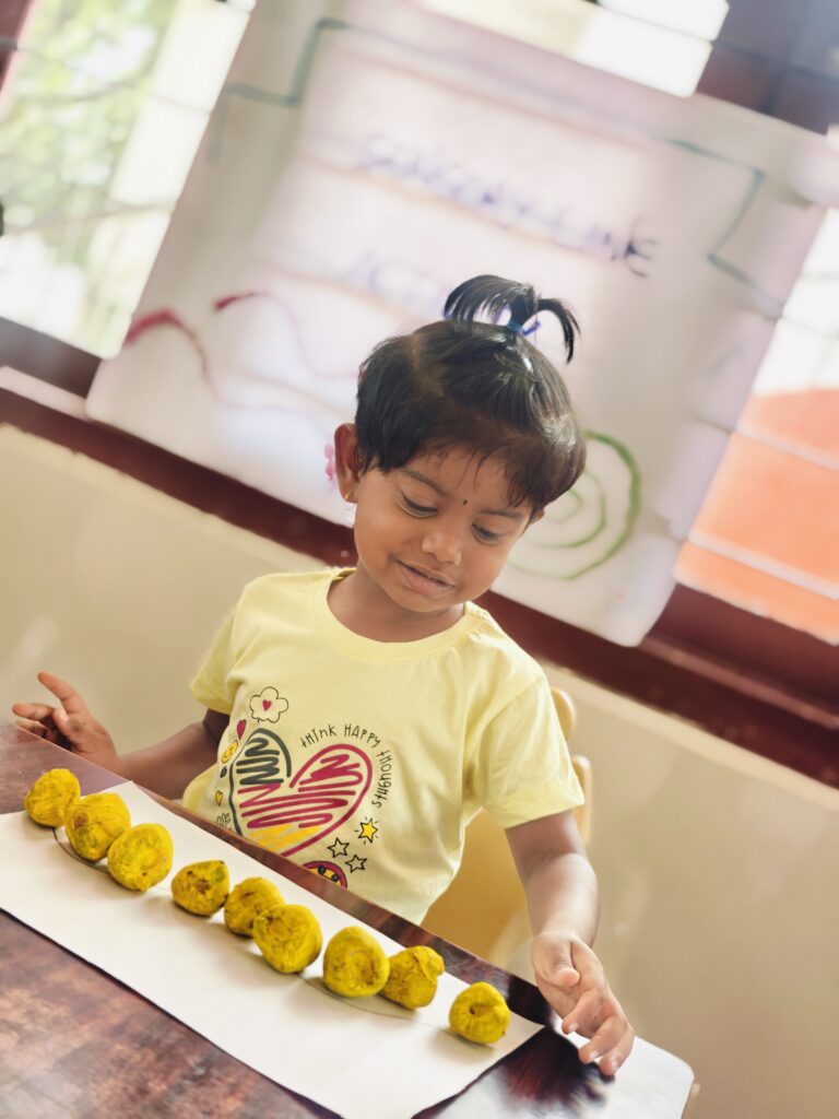 Child with a top knot smiles while placing yellow pieces along a long line on paper.