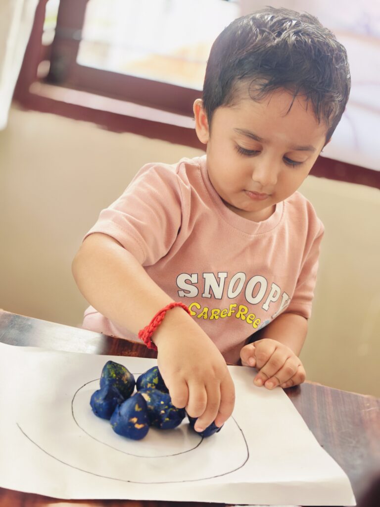 Child in a pink T-shirt places dark blue pieces along a drawn circular line on white paper.