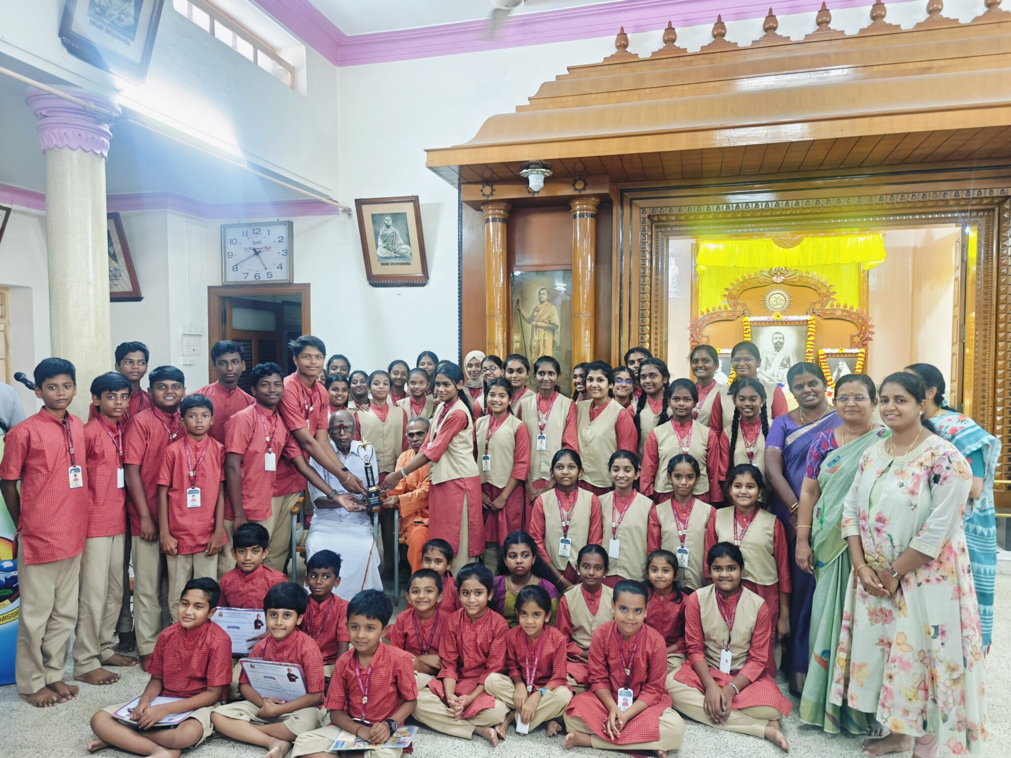 Students in red uniforms with the National Youth Day Competition trophy in a hall, with staff and seated guests
