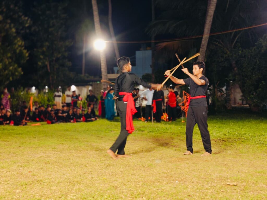 Two students in black with red sashes cross wooden sticks during a sparring sequence
