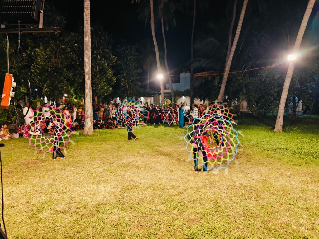 Three performers spin large multicoloured circular frames on a lawn as an audience watches