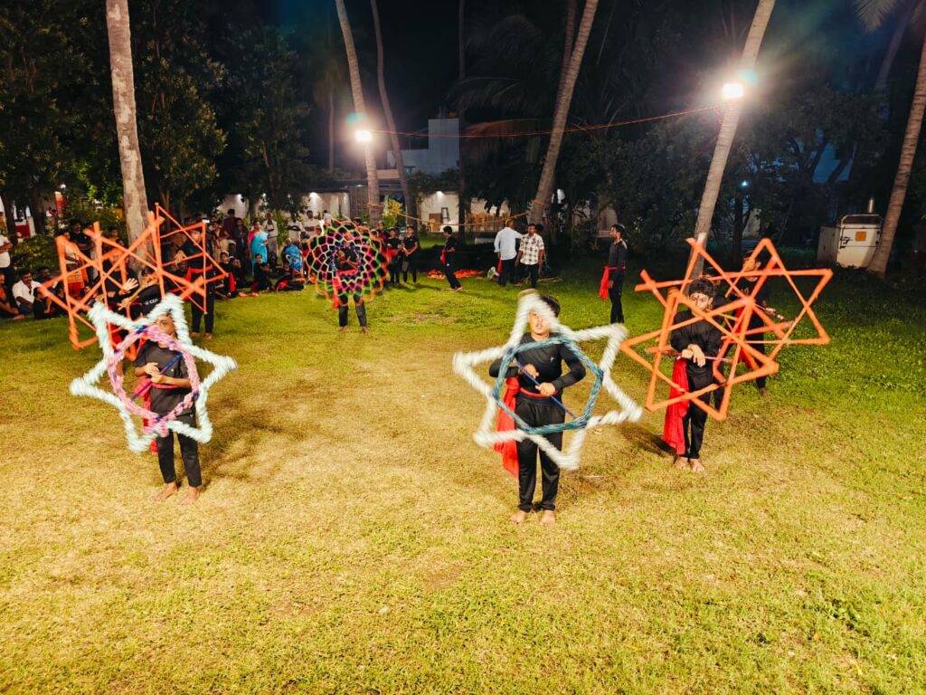 Four students spin large white, blue, pink and orange star-shaped frames on a floodlit lawn at night