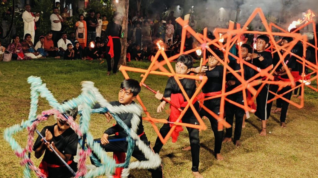 Silambam performance: students in black and red spin star-shaped frames as torches burn and an audience watches