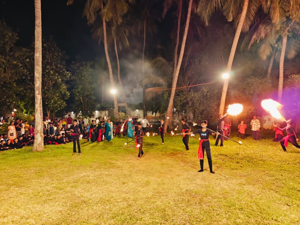 Students twirl staffs with flames at both ends as smoke rises and an audience looks on