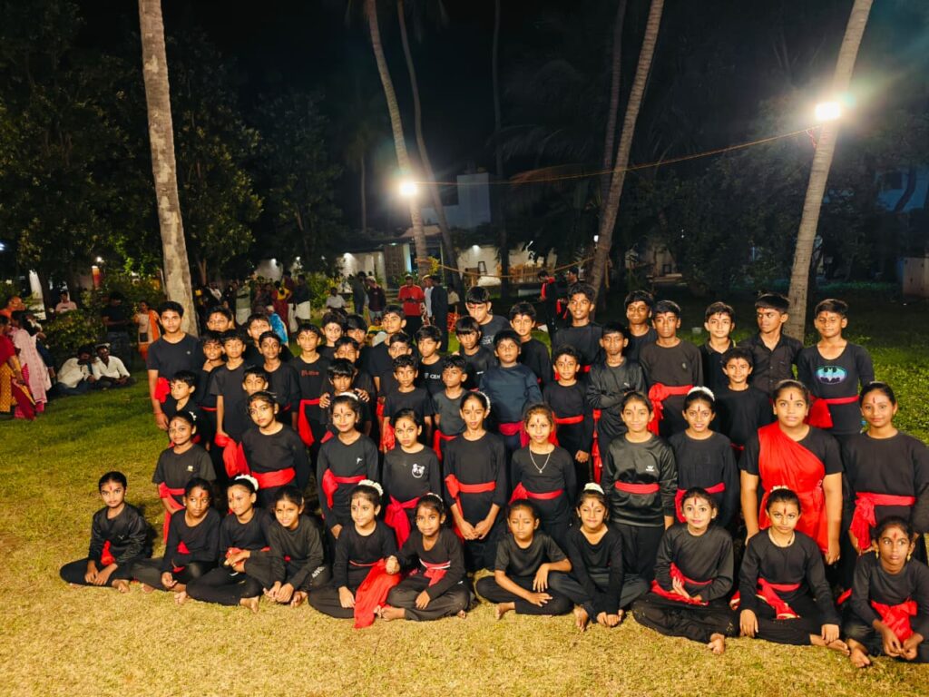 Group photograph of the performers in black outfits with red sashes, seated and standing on the grass