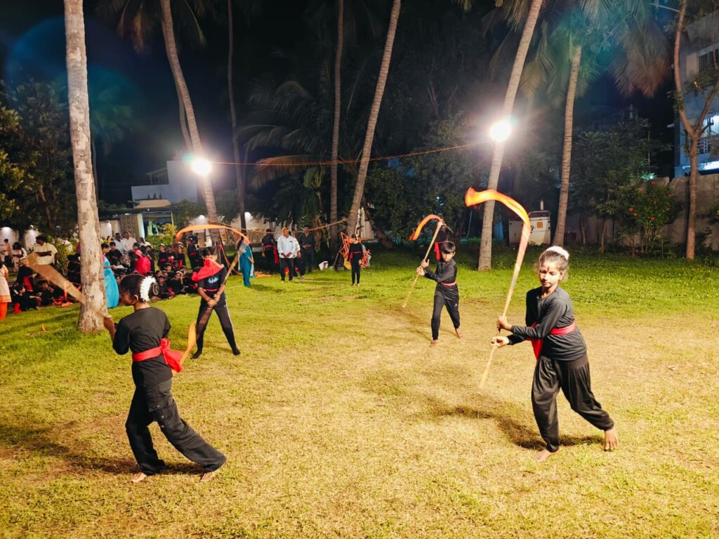 Students swing sticks trailing orange ribbons on a floodlit lawn among palm trees