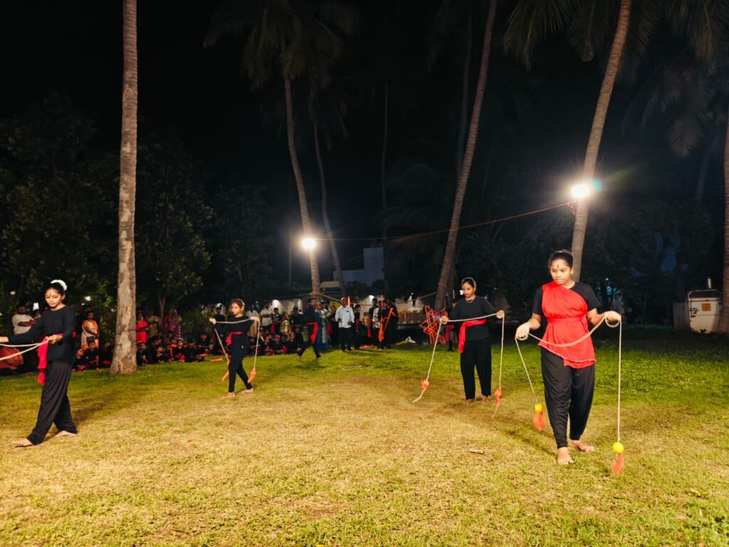 Four girls perform with long ropes ending in weighted orange tassels on a floodlit lawn