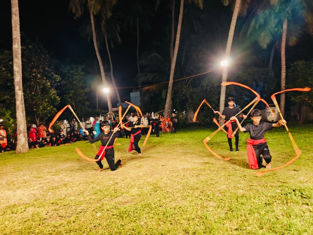Boys kneel and swing sticks with long orange ribbons on the grass at night