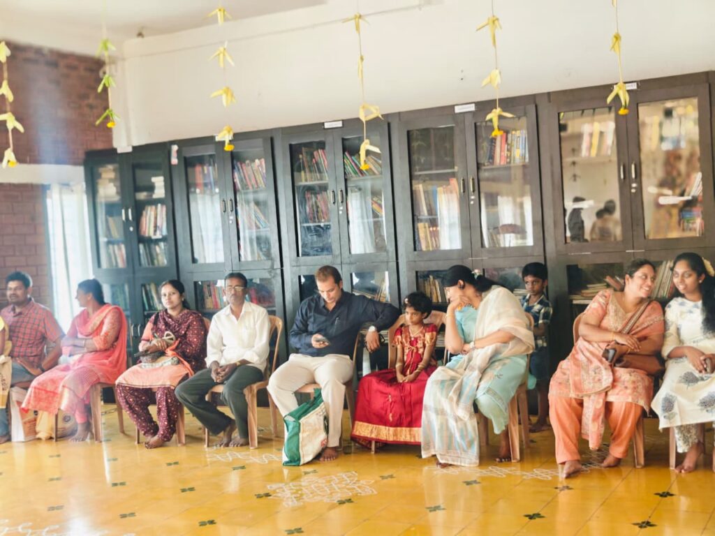 Adults and children seated on chairs before glass-fronted bookcases, with palm-leaf decorations hanging above.