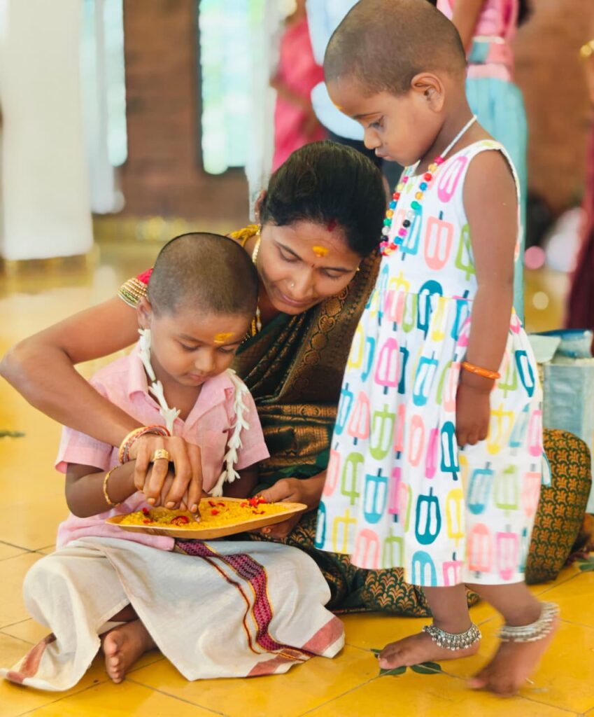 A woman guides a child's hand in a tray of rice while a small girl in a printed dress stands watching.