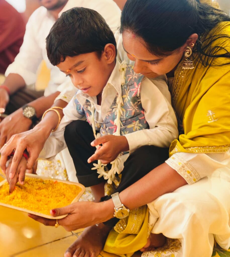 A woman in yellow guides a boy's finger through a tray of turmeric rice on the floor.