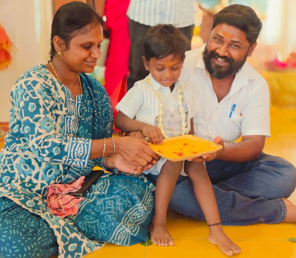 A boy wearing a jasmine garland touches a tray of turmeric rice held by two adults sitting on the floor.