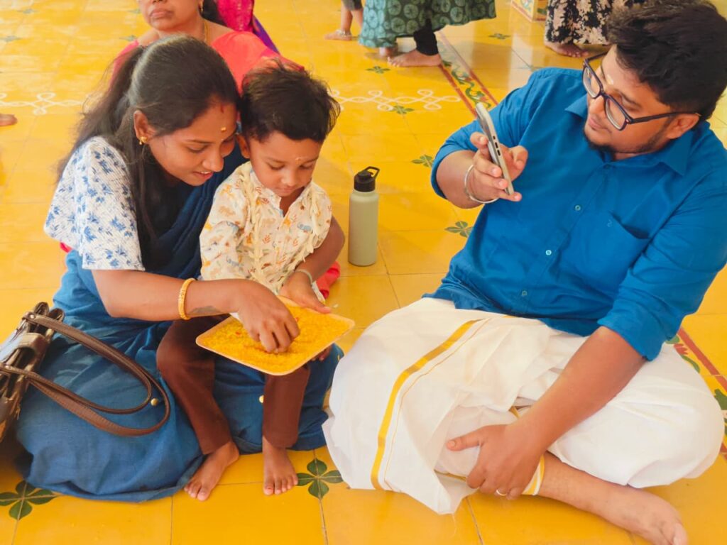 A woman guides a toddler's hand in a tray of yellow rice as a man in a veshti photographs them.