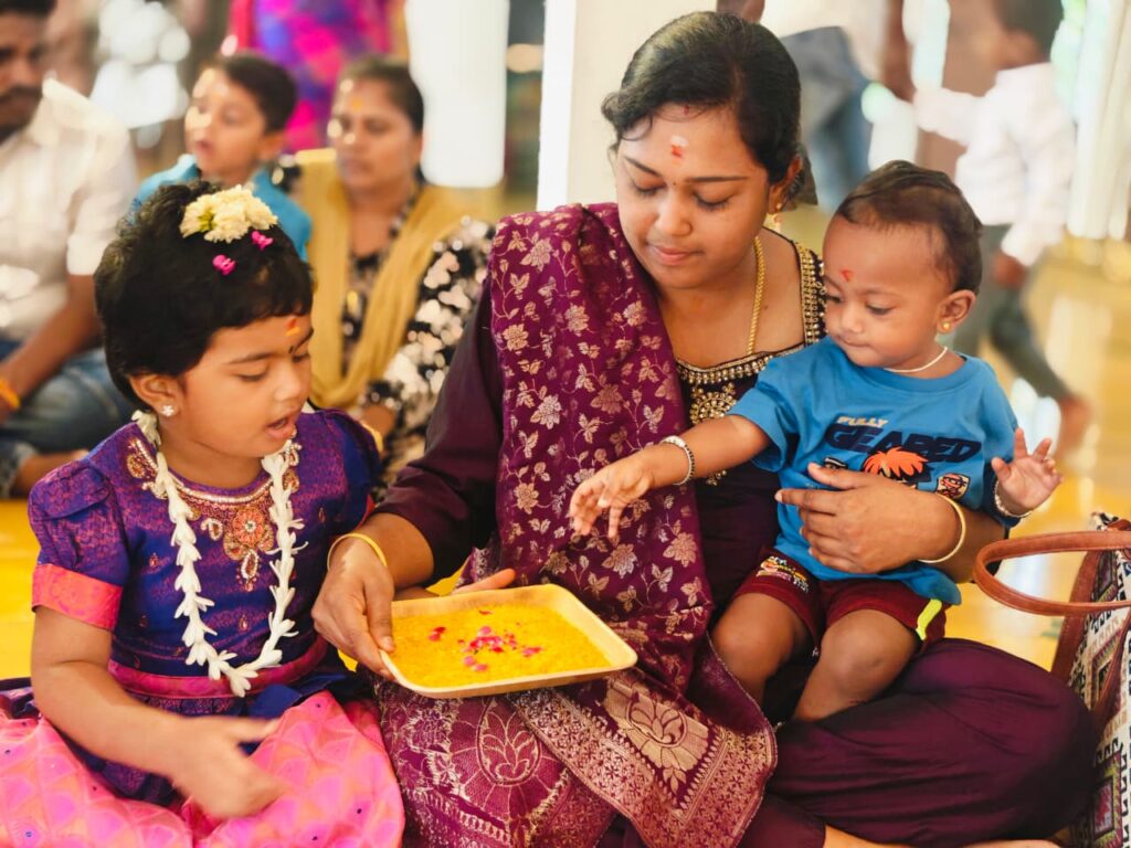 A woman holds a tray of yellow rice as a toddler on her lap reaches towards it and a girl in purple watches.