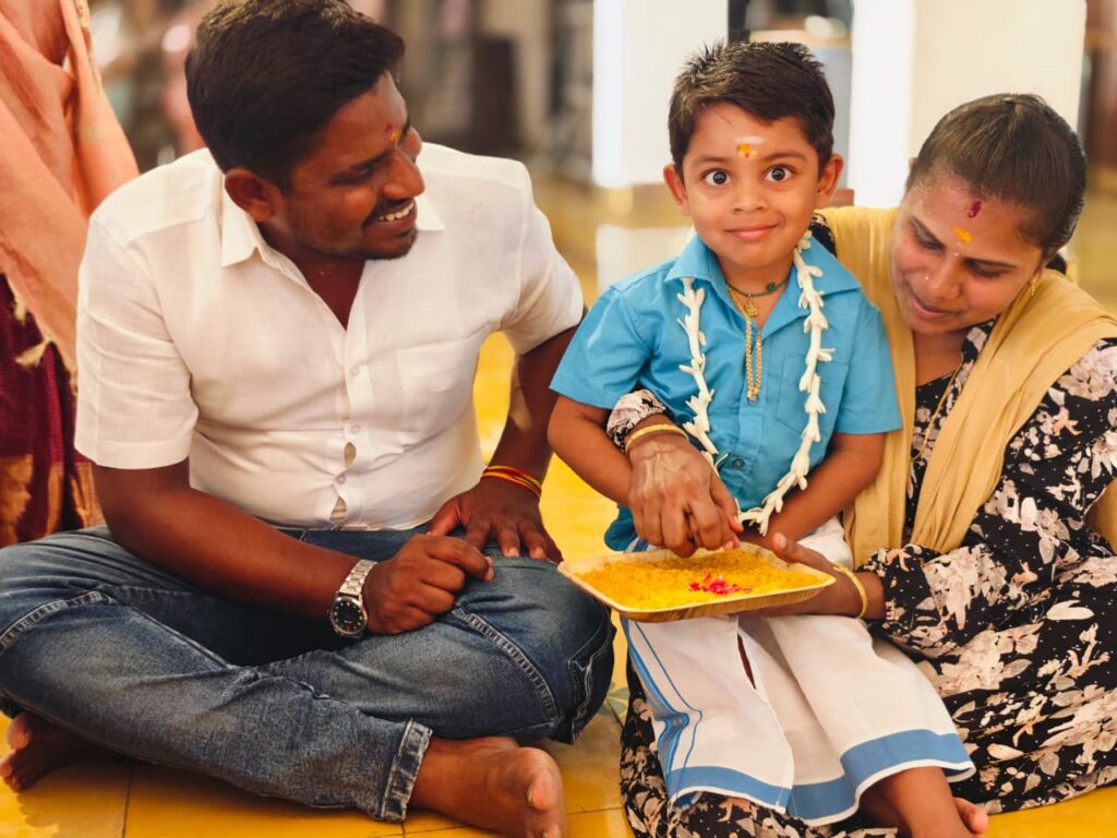 A boy in a blue shirt and garland smiles as an adult guides his finger through a tray of yellow rice.