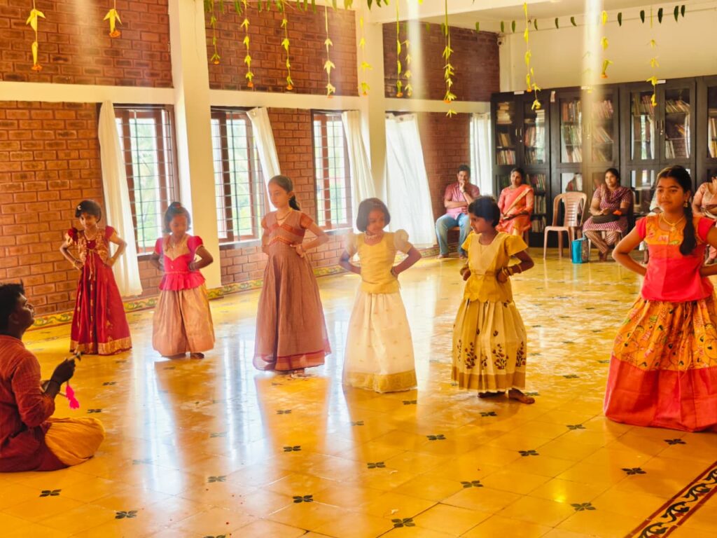 Girls in long traditional skirts dance in a decorated hall while a man sits on the floor and adults watch.