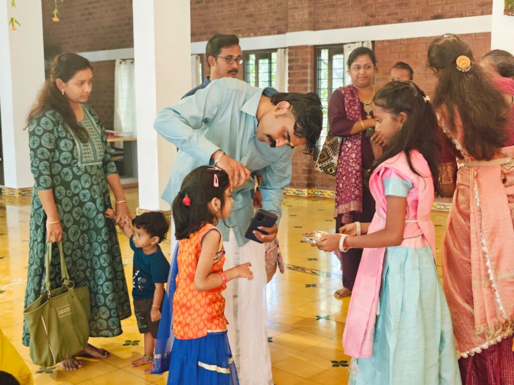 A man places a mark on a young girl's forehead while an older girl holds a small plate and adults look on.