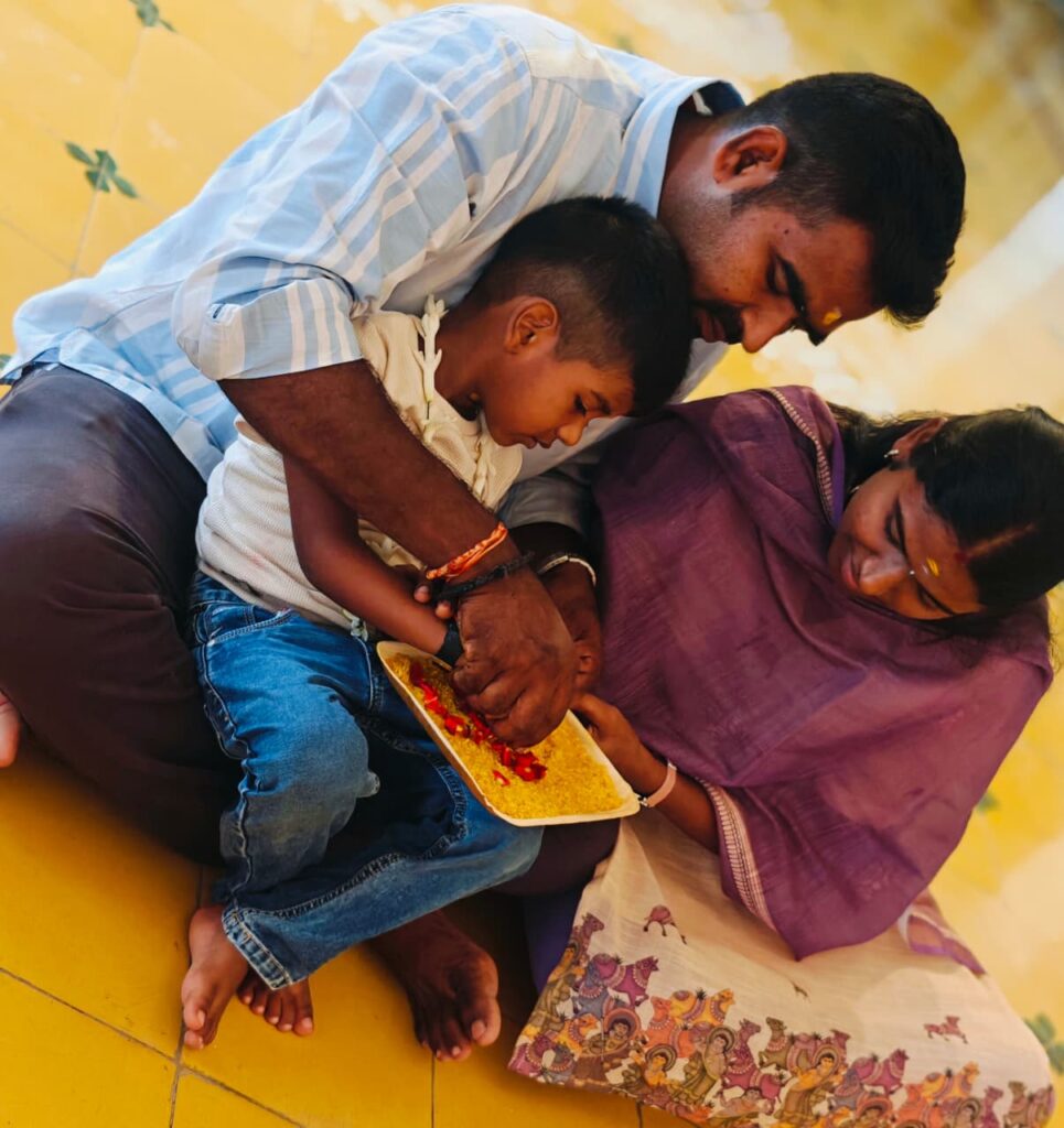 A man and a woman lean over a boy as he traces in a tray of yellow rice with red petals.