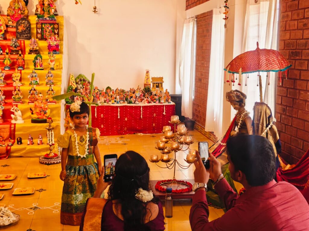 Kindergarten Aksharaabhyaasam: a child in a silk pavadai stands before a doll display as two adults take photos.